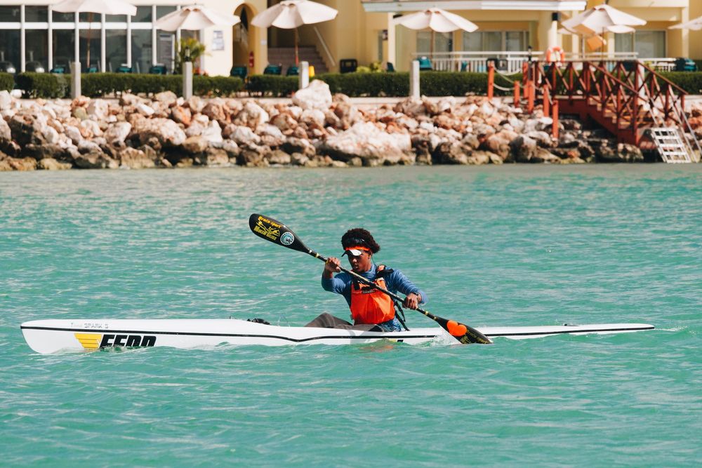 A kayaker in red life vest paddles a white kayak across turquoise waters near a rocky shoreline.