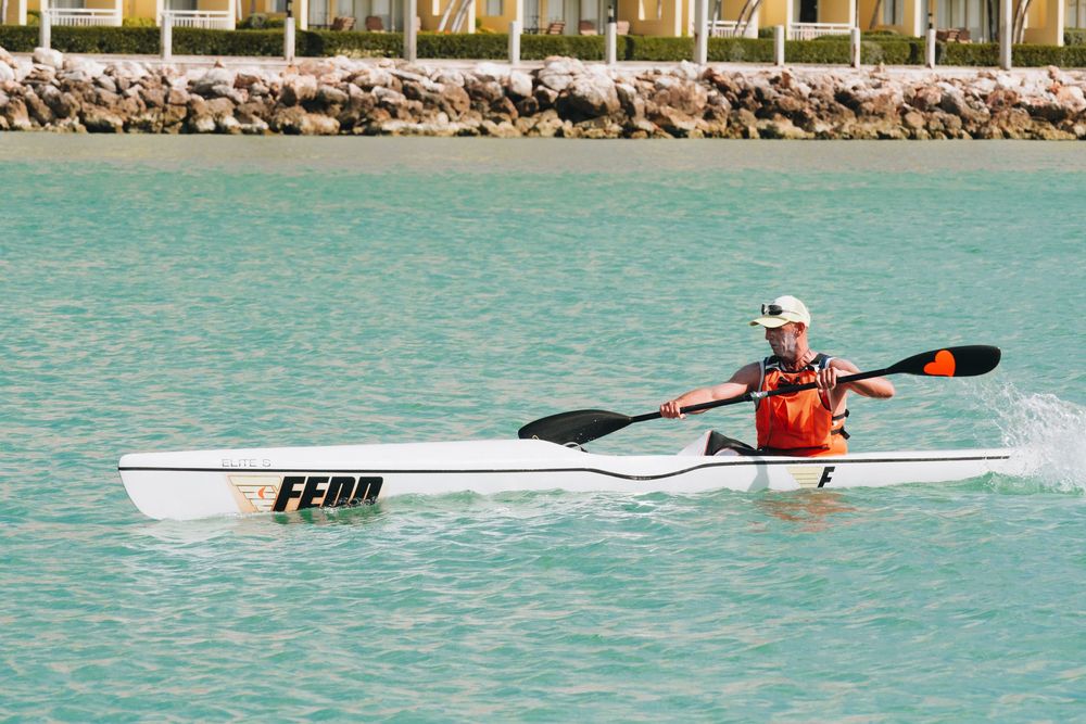 A kayaker paddling a white racing kayak across turquoise waters near a rocky shoreline.