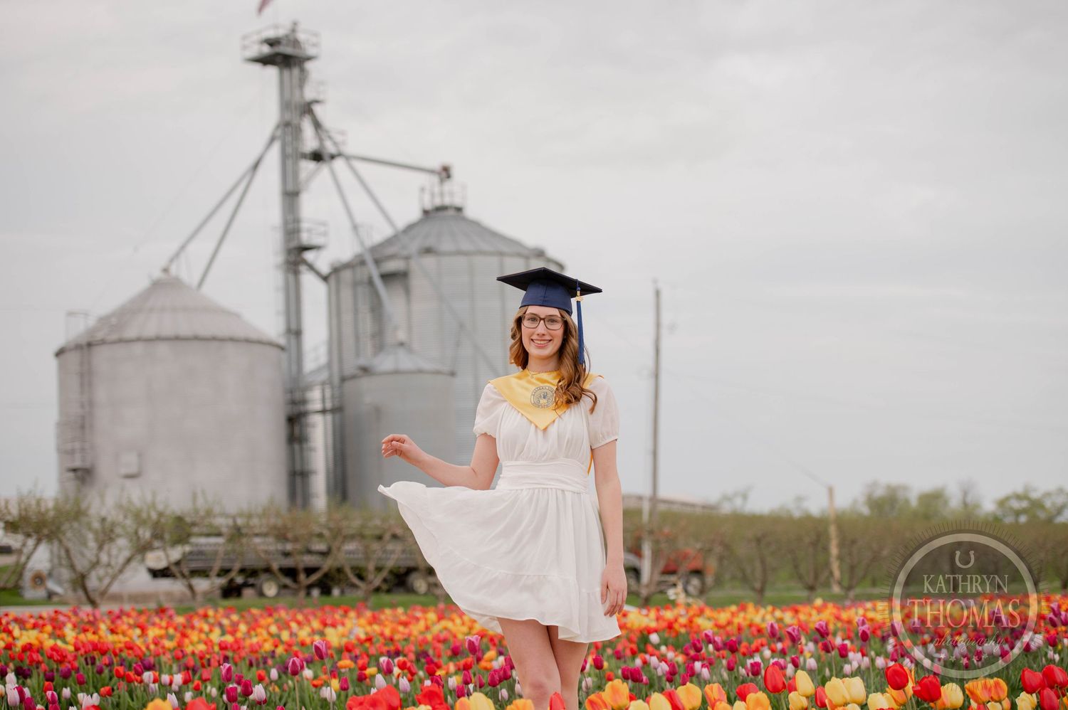 A graduate poses in a flowing dress among vibrant tulip fields in front of industrial grain silos on a cloudy day.