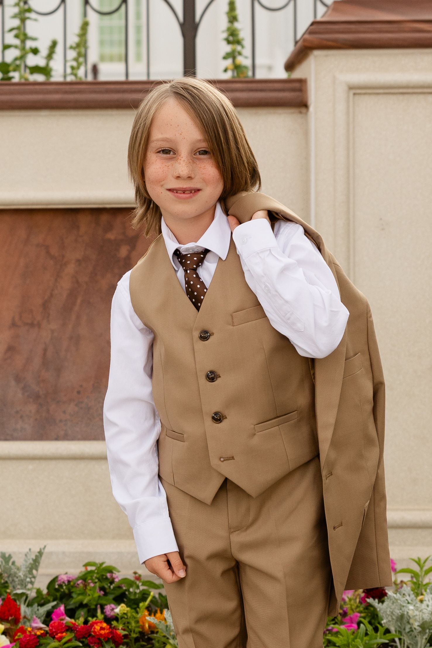 8 year old boy in brown 3 piece suit smiling at the camera. His suit jacket flung over one shoulder