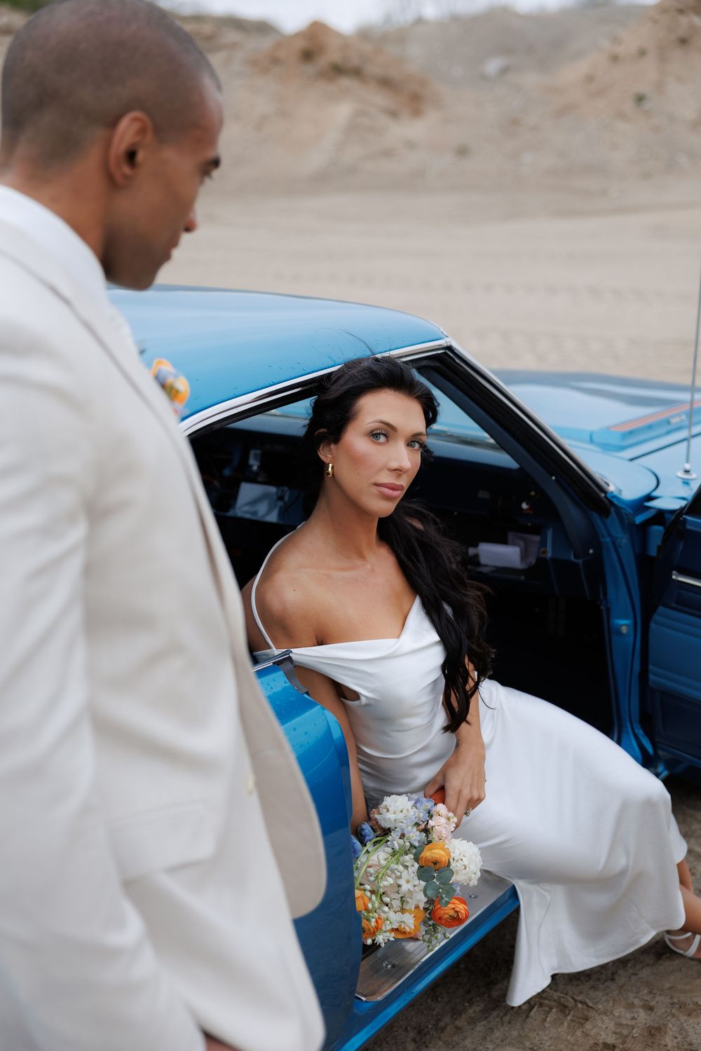 Wedding moment captured as couple interacts beside classic blue vintage car.