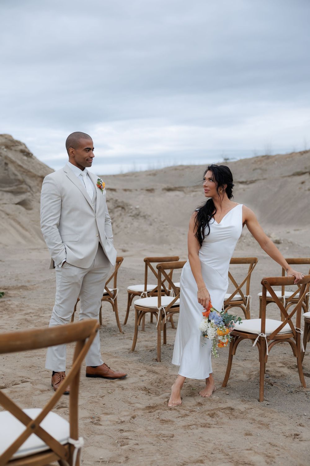 Desert wedding scene with couple in white attire posing among wooden chairs.