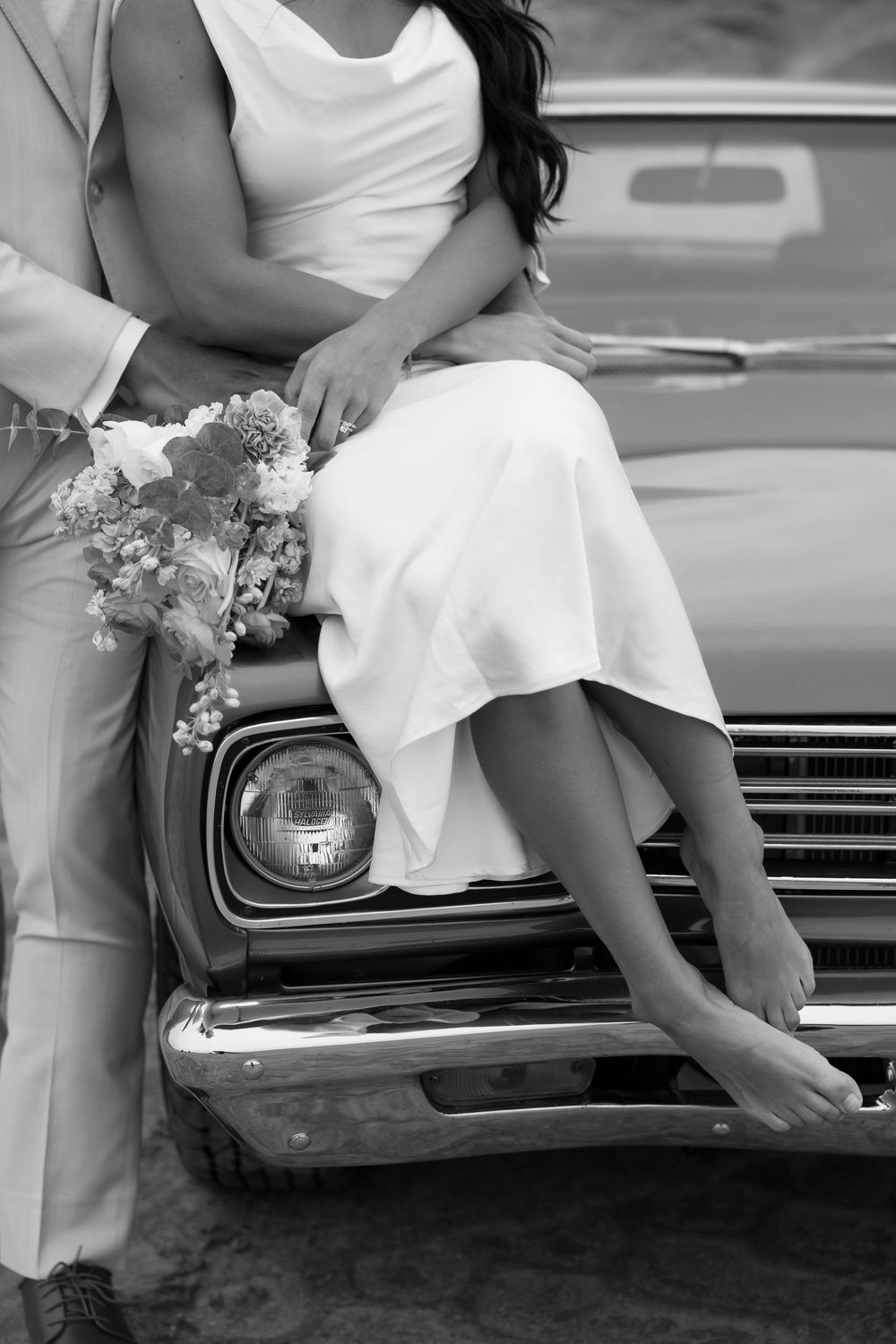 Couple in white wedding attire sitting on vintage car hood with small floral bouquet.