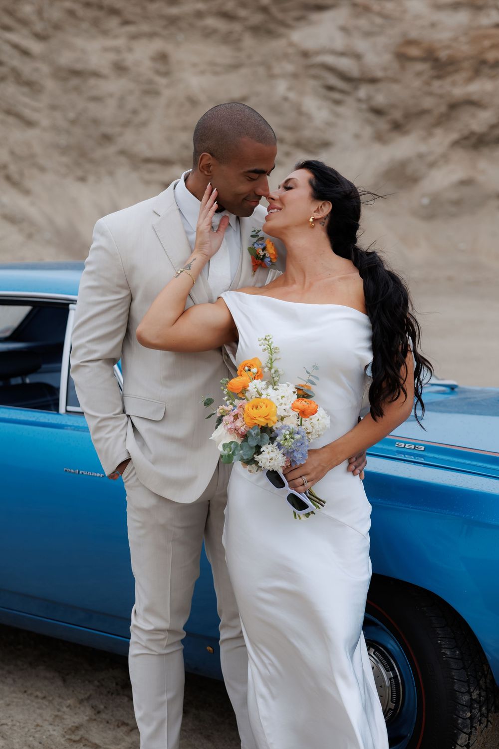 Romantic wedding couple embracing near a vintage blue car in a desert landscape.