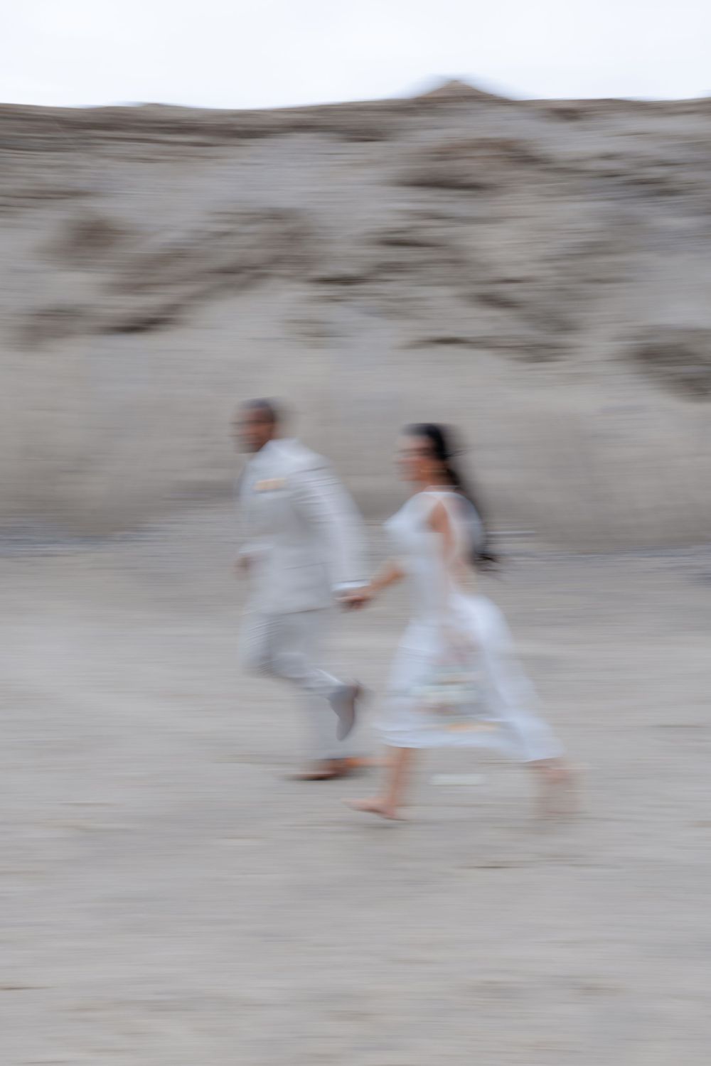 Motion-blurred photograph of couple in white attire walking on desert sand.