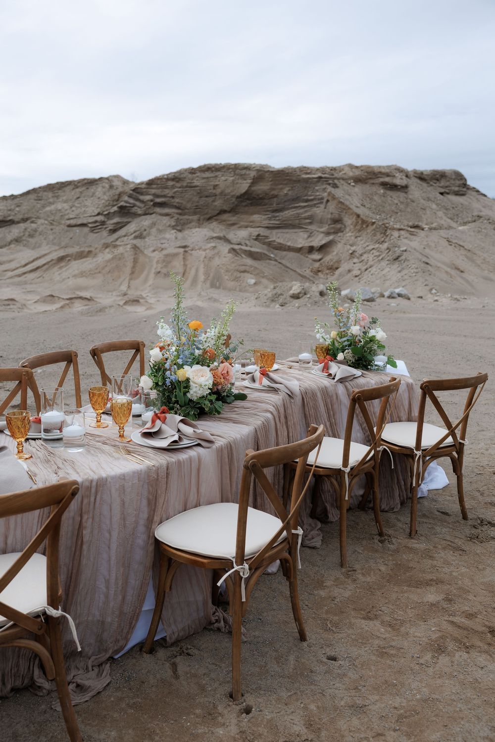 Rustic outdoor wedding reception table set with wooden chairs and delicate floral arrangements.