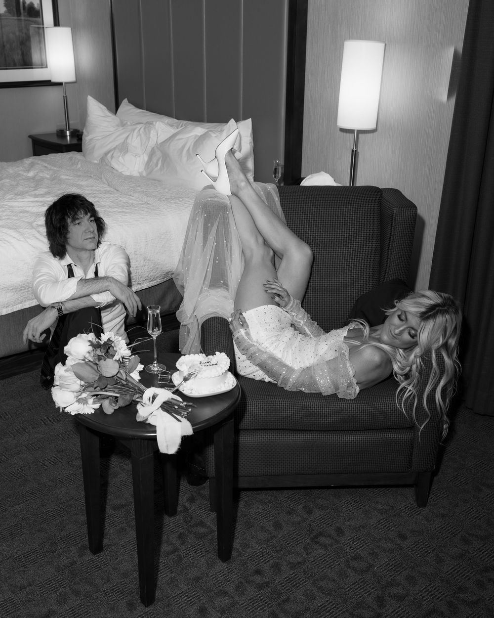 A black and white photo of newlyweds relaxing with cake and champagne in a hotel room.