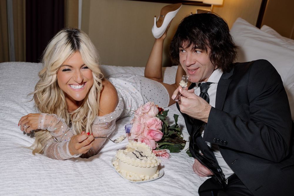 A newlywed couple lying on a bed sharing cake and laughter during their wedding celebration.