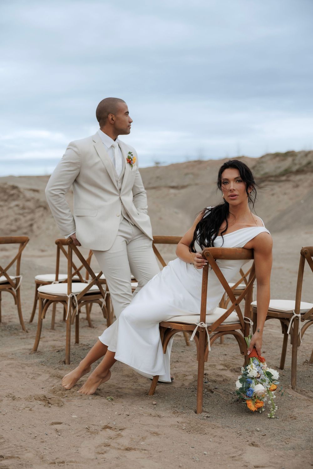 Elegant beach wedding scene with wooden chairs and a bridal bouquet on sandy terrain against cloudy sky.