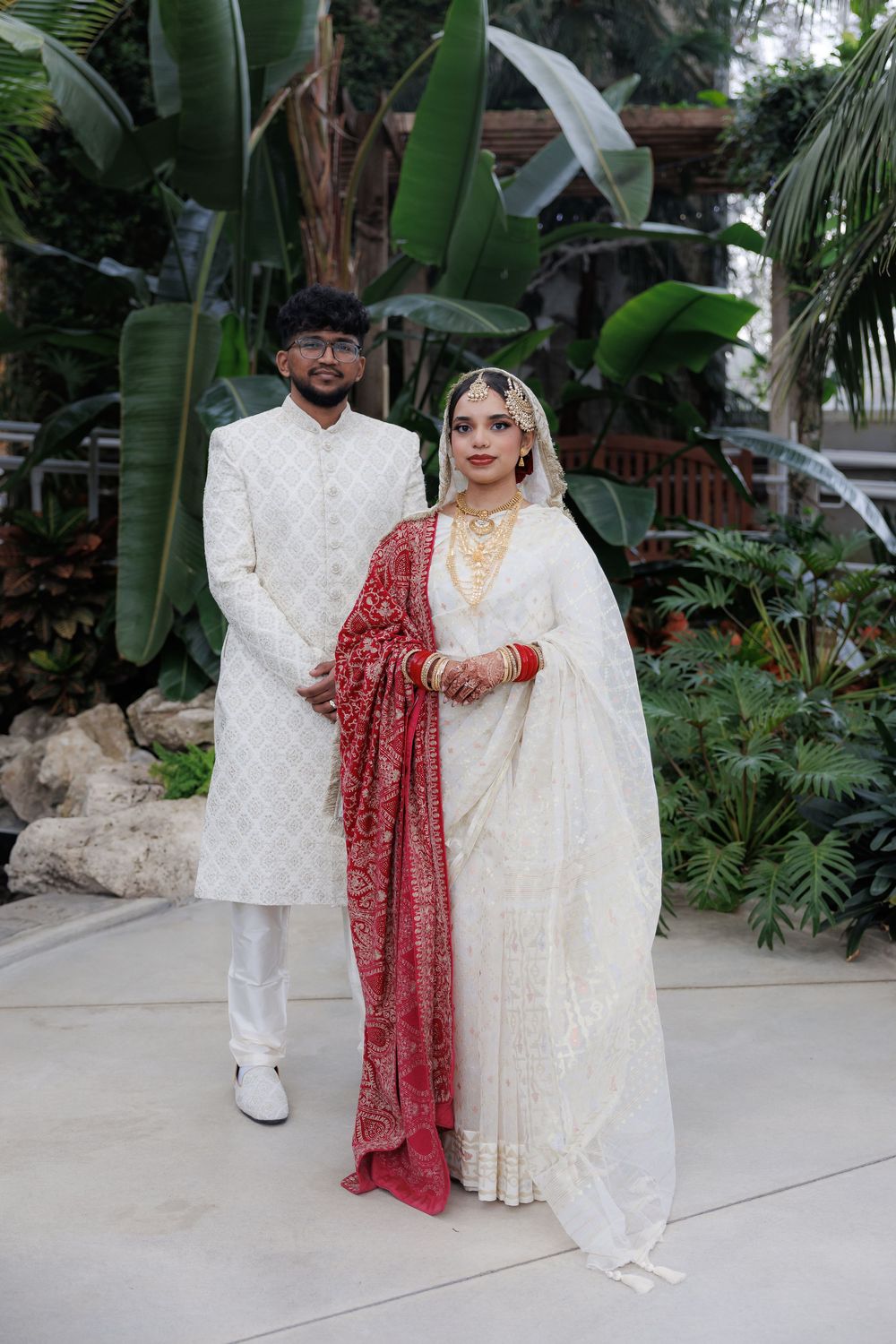 Wedding couple in traditional white and red attire posed in a lush tropical garden setting.