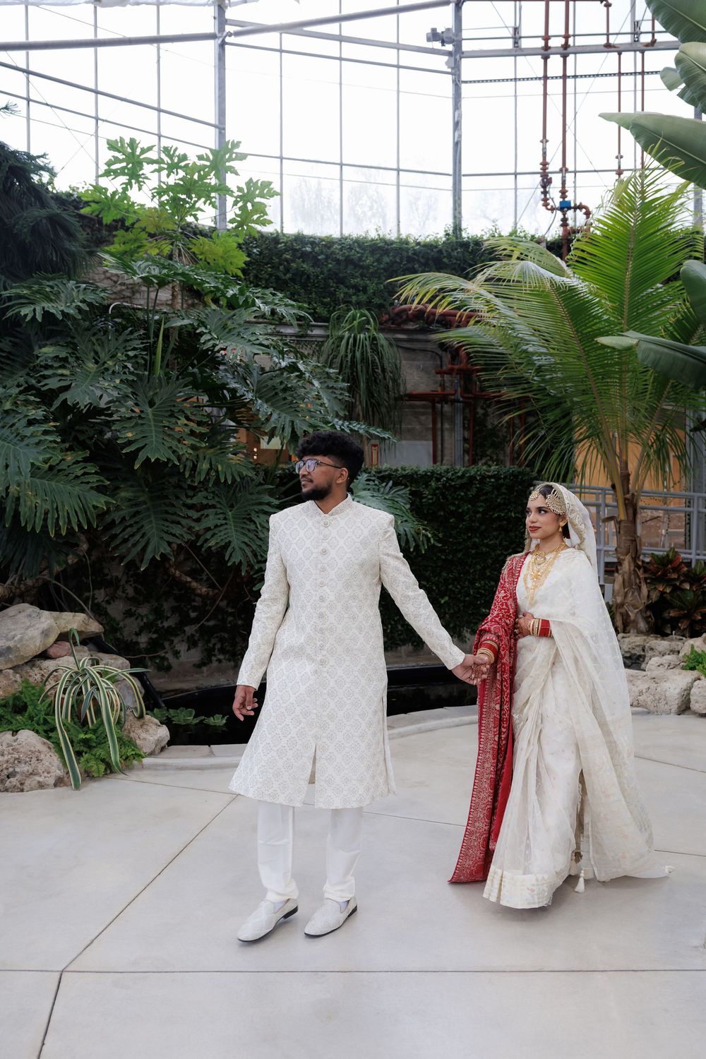 A couple in traditional South Asian wedding attire standing in a greenhouse setting.