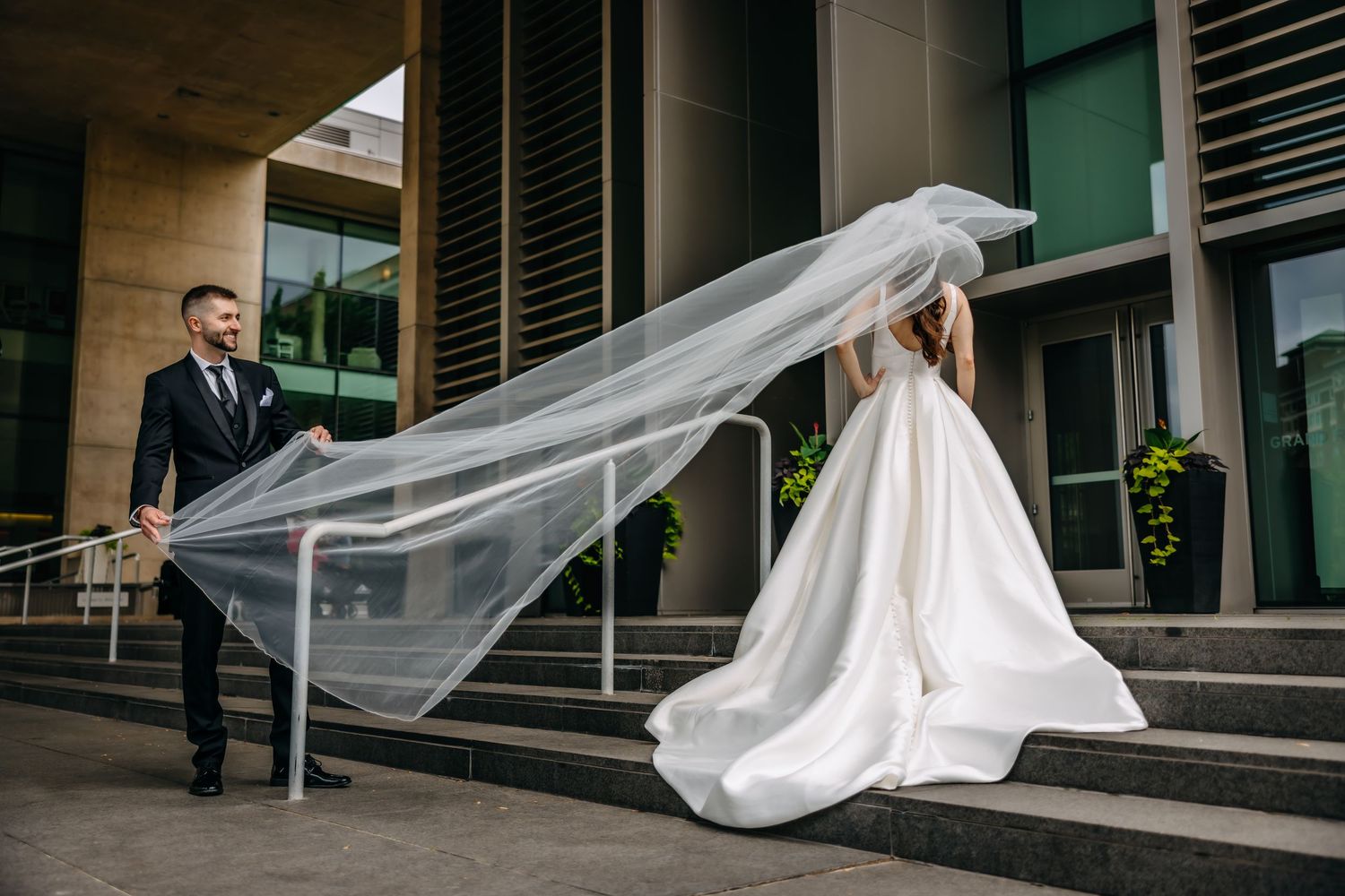 A dramatic wedding photo with flowing white veil catching the wind on modern building steps.