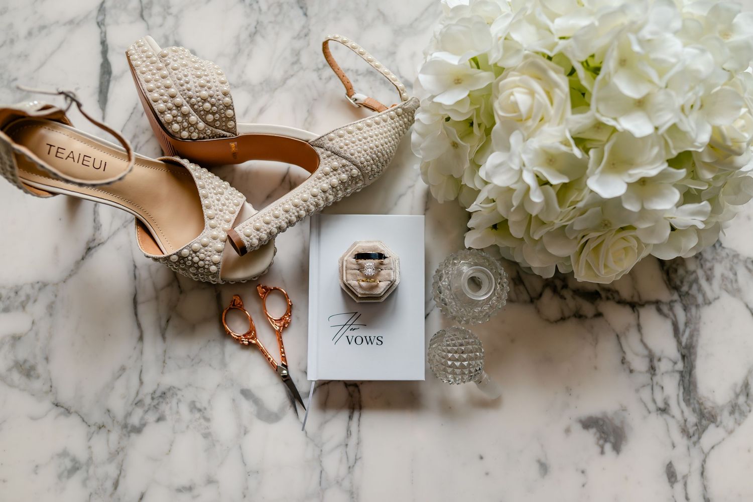 Wedding accessories including bridal shoes, perfume bottle, white hydrangea bouquet, and jewelry on marble surface.