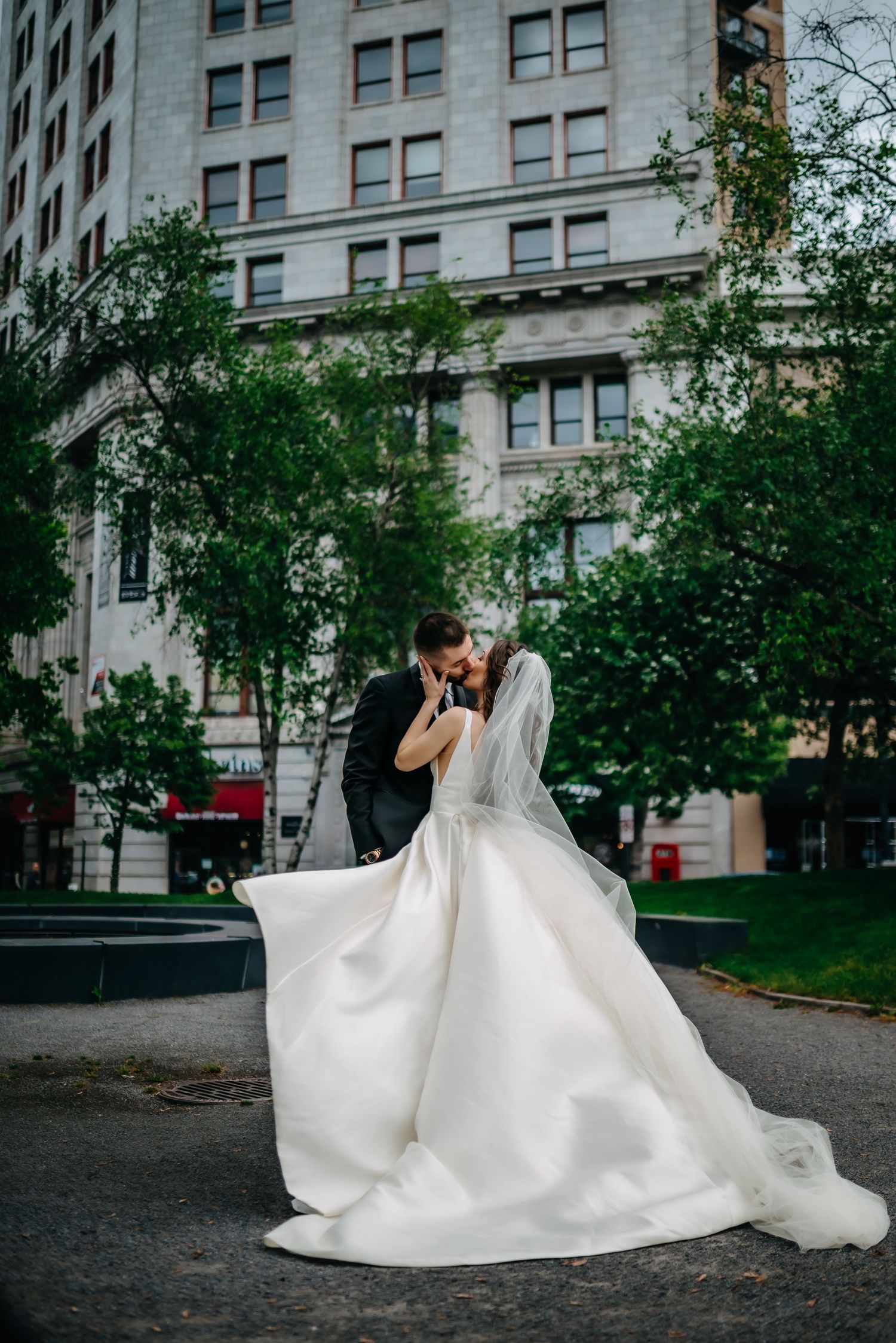 A wedding couple embraces in a romantic kiss against an urban backdrop with trees and a grand building.