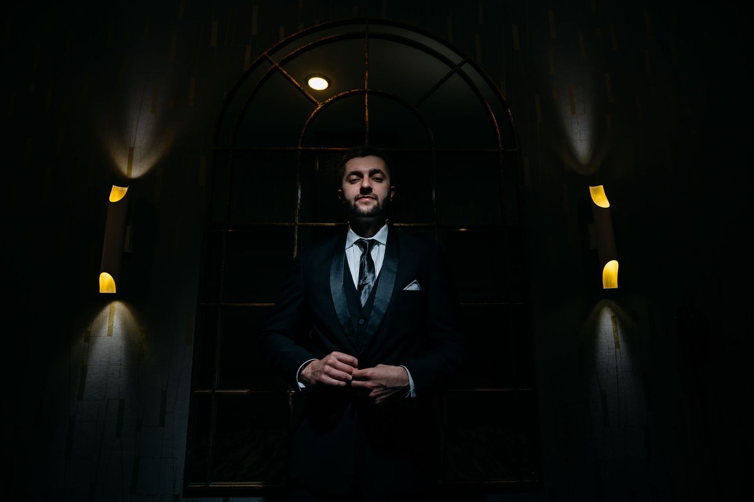 Moody lighting illuminates figure in dark suit against arched doorway with architectural details visible.
