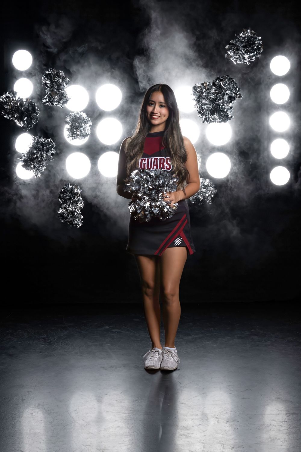 Cheerleader poses with silver pom poms against dramatic backdrop with bright spotlights and smoke effects.