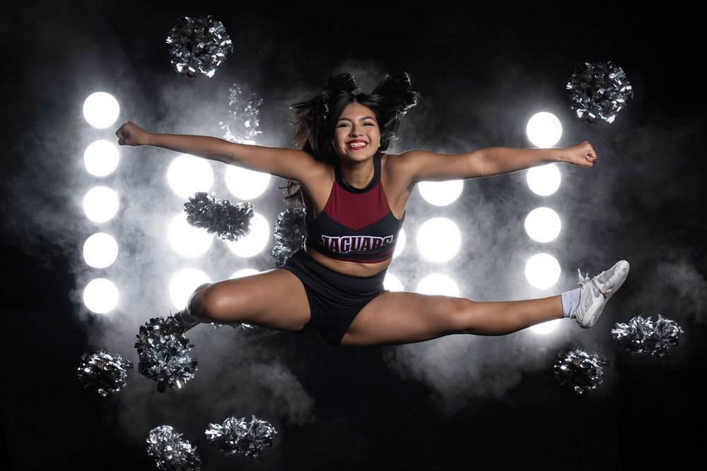 Athletic cheerleader performs energetic toe touch jump against dramatic spotlit background with smoky effects.