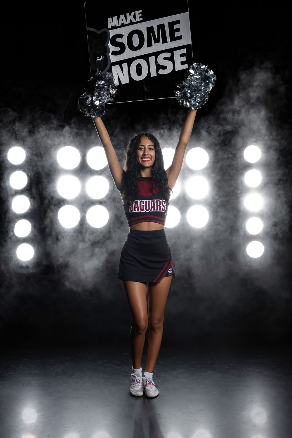A cheerleader in black uniform holds up a 'MAKE SOME NOISE' sign with pom poms against bright stage lights and smoke effects.