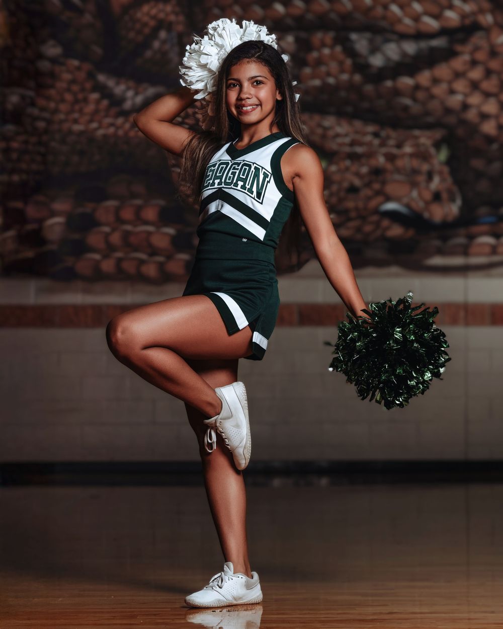 A cheerleader in a green uniform strikes a dynamic pose with pom poms against a brick wall backdrop.