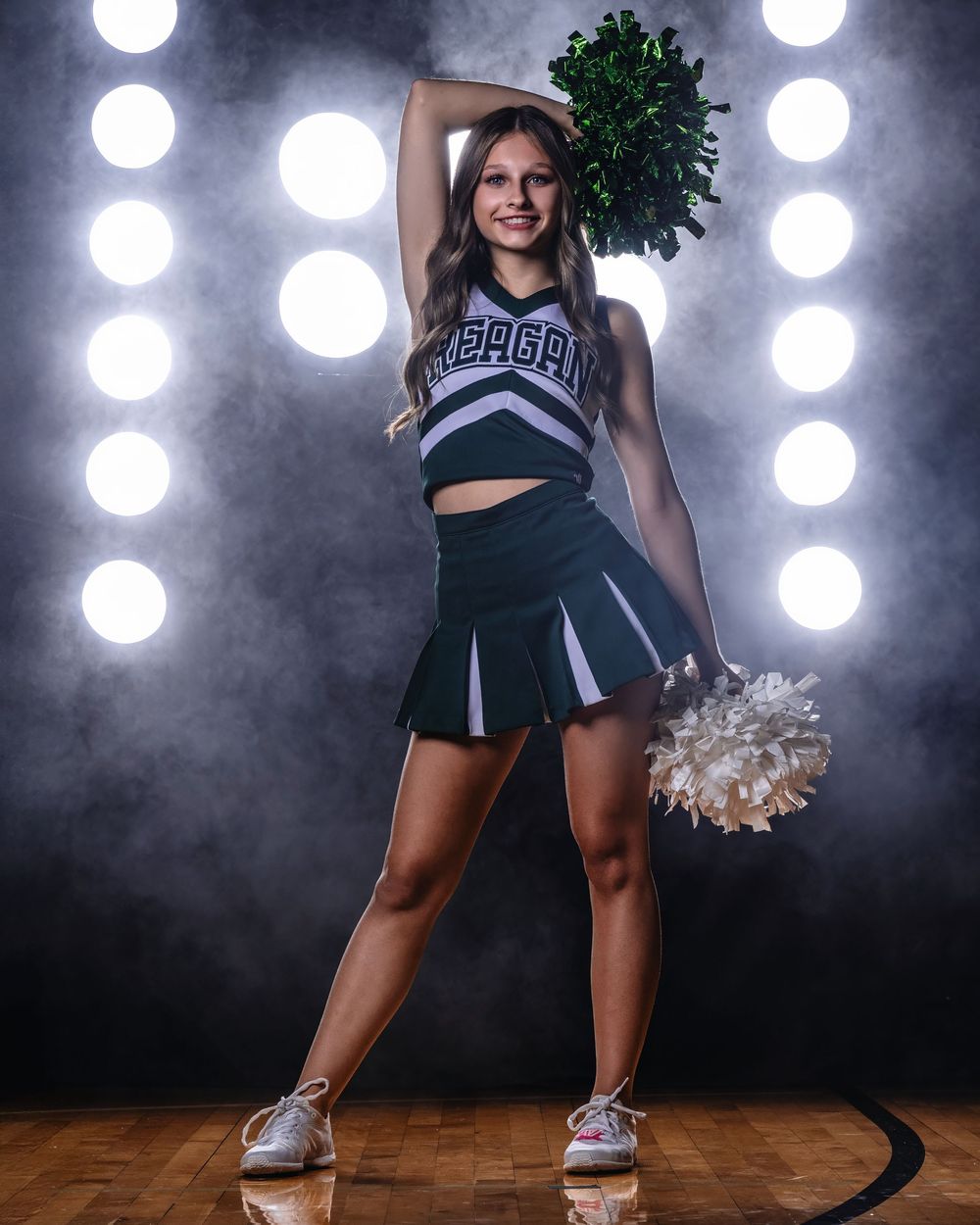 A cheerleader in navy and white uniform performs with pom poms against a dramatic backdrop with spotlights.