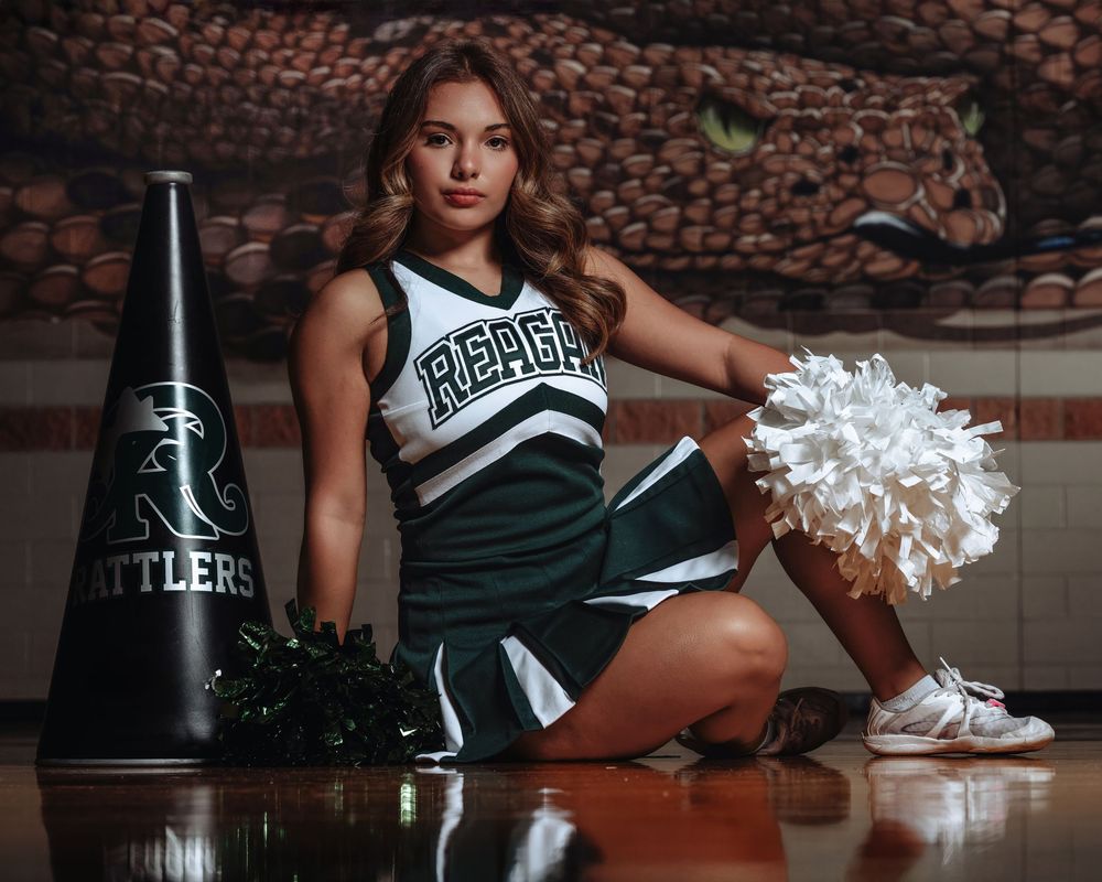 Cheerleader in green and white uniform poses with pom poms on brown gymnasium floor with dramatic lighting.