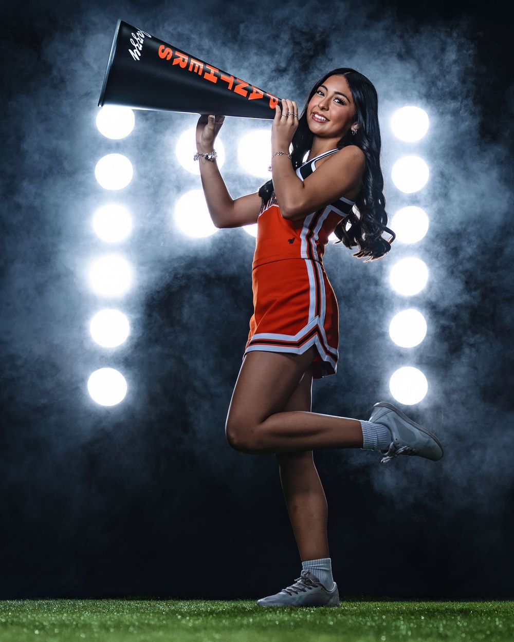 Cheerleader in orange uniform performs dynamic jump pose against dramatic dark background with bright spotlights.