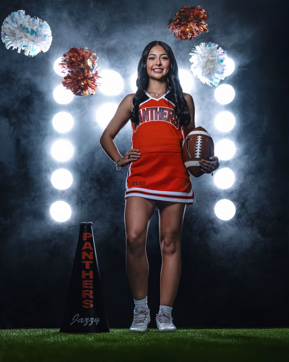 A student athlete in a red and white cheerleading uniform poses with a football against a dramatic backlit backdrop.