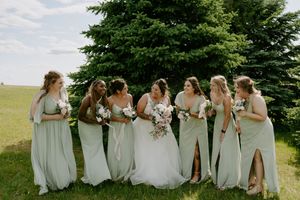 Bridal party in sage green dresses sitting together on grassy hillside.