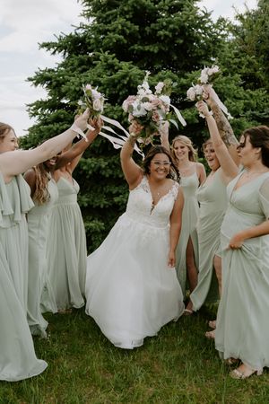 Joyful bridal party in sage green dresses celebrating outdoors with raised arms.