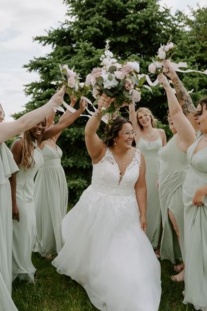 Wedding party in sage green dresses and white gown pose playfully outdoors with raised arms.
