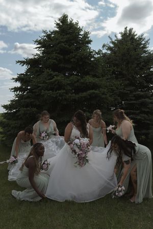 Bridal party in sage dresses sits together on grassy field with evergreen trees.