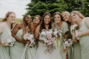 Bridesmaids in sage green dresses share joyful moment with bouquets outdoors.