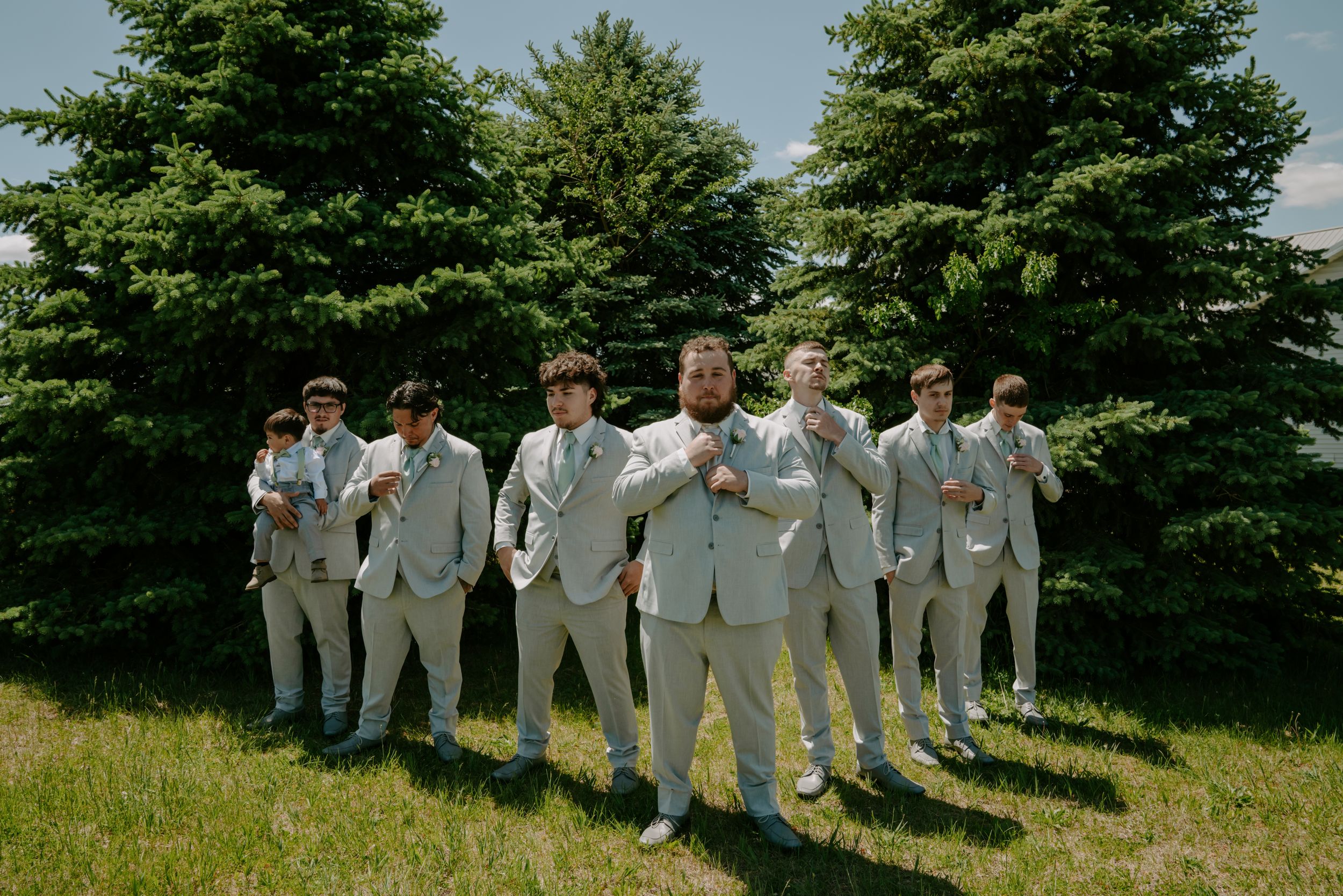 Wedding party in light gray suits standing together on grassy hill with evergreen trees.