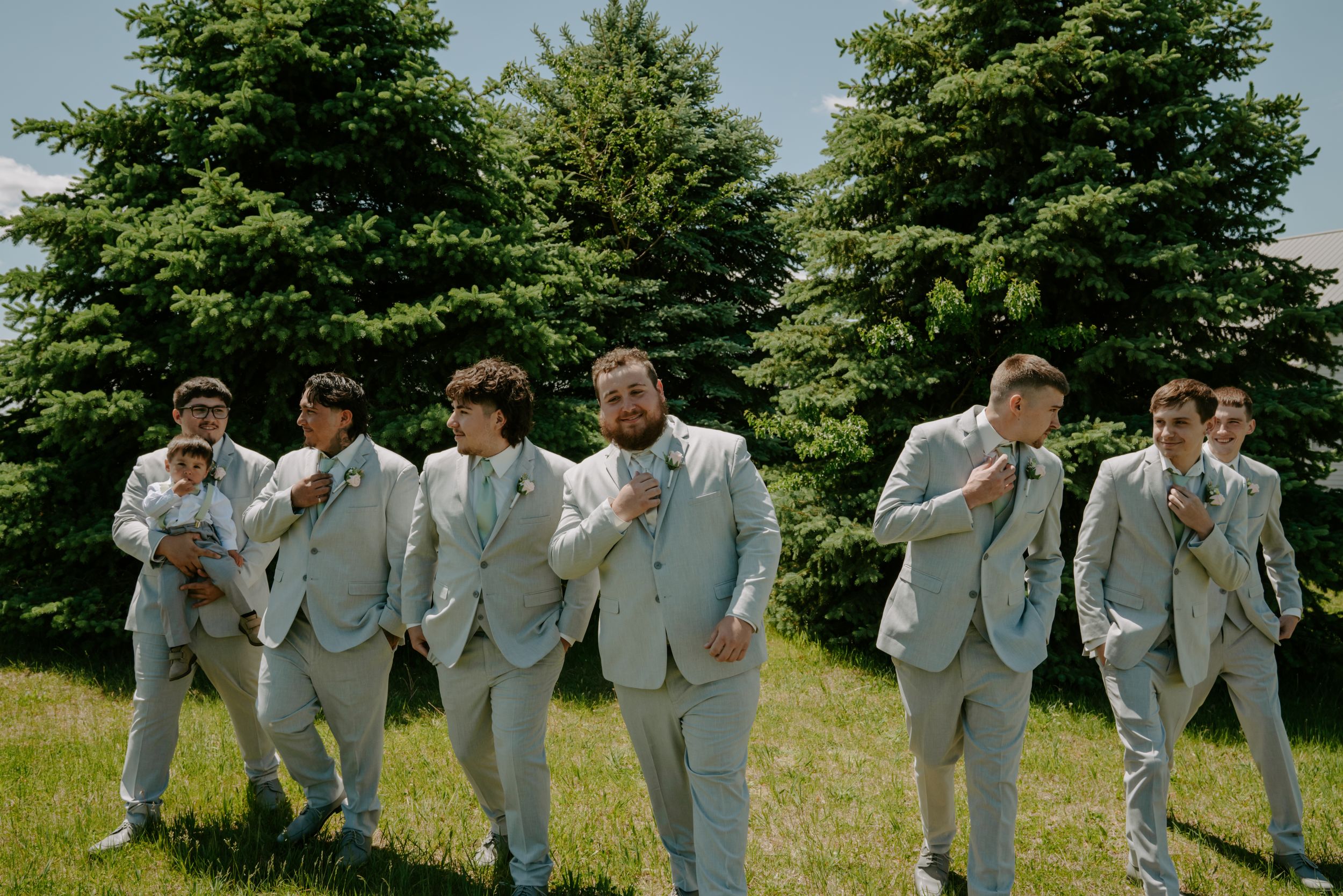 Groomsmen in light gray suits pose playfully in a line on a sunny day.
