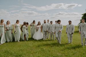 Large wedding party in white and light gray attire poses in line on grassy hill.