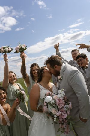 Wedding party celebrates joyfully outdoors with bouquets of pink and purple flowers against a blue sky.