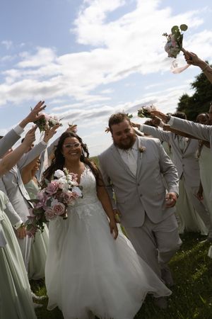 Wedding party jumps and celebrates together on grassy hillside with flowers and formal attire.