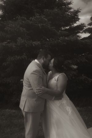 A romantic wedding couple shares a tender kiss outdoors with trees in the background.