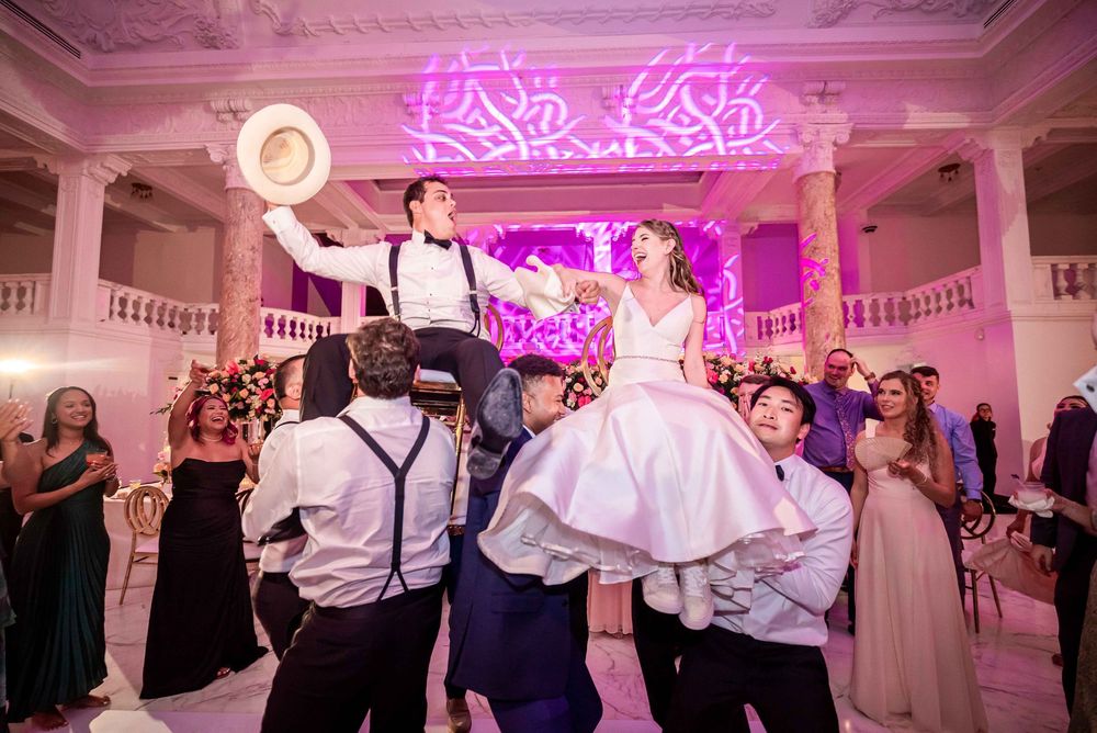 Guests dance and celebrate at a wedding reception venue with pink neon sign lighting and elegant white decor.