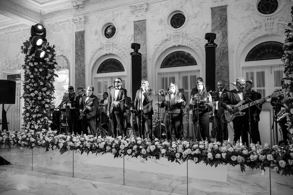 Black and white photo of a band performing on stage with floral decorations at an elegant venue with arched windows.