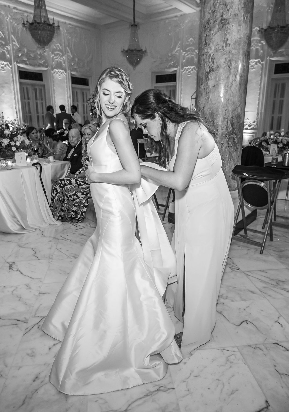 Black and white photo of a wedding dress being adjusted in an ornate marble ballroom with chandeliers.