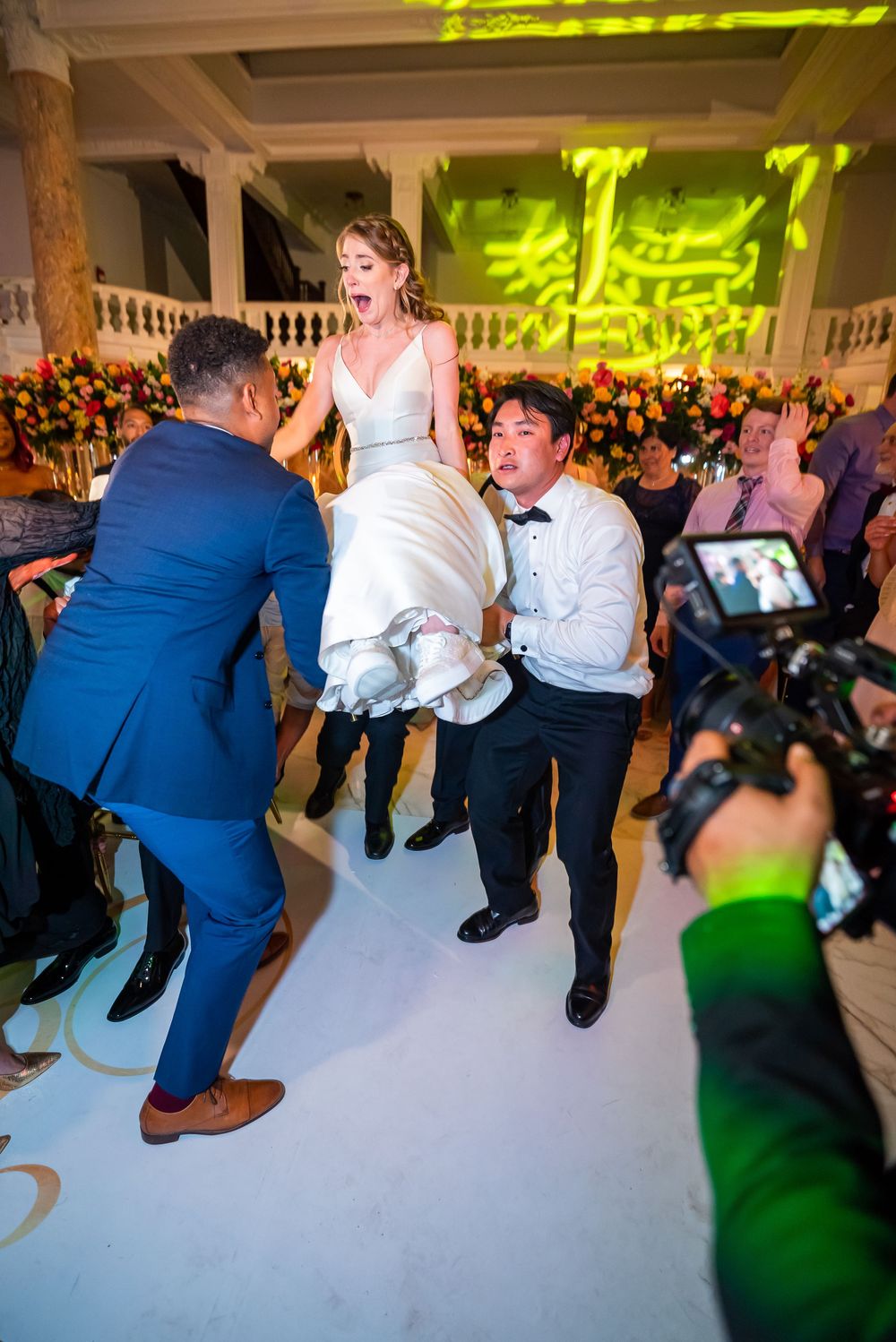 Wedding guests lift bride while dancing on white floor at reception with green lighting effects.