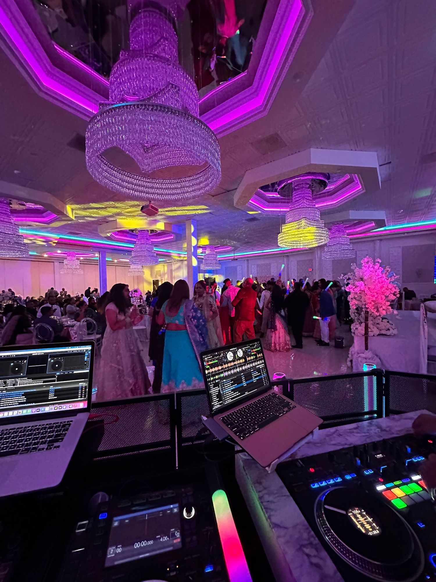 A DJ's station overlooks a crowded formal event with purple lighting, crystal chandeliers and colorful ceiling accents.