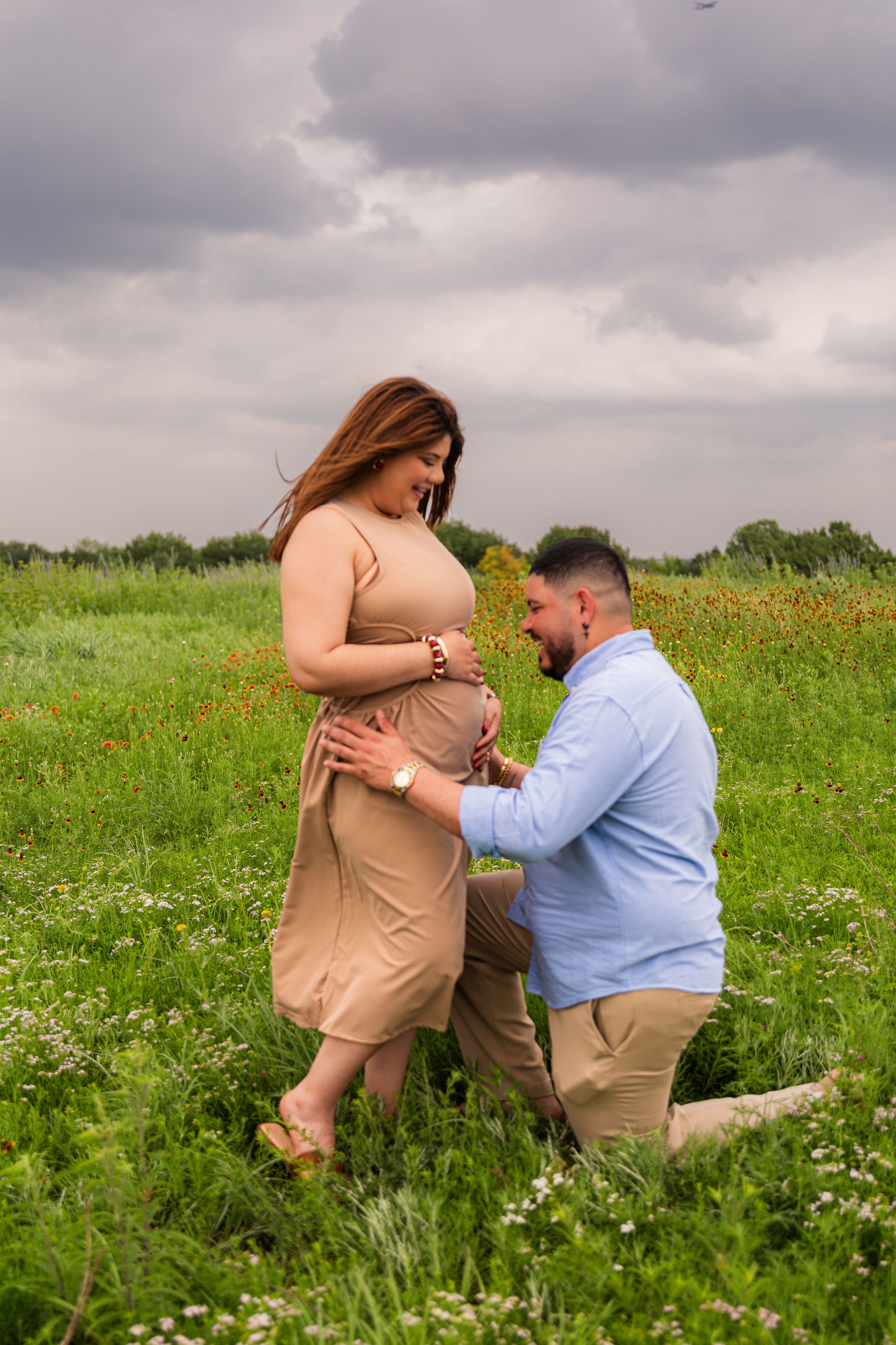 A man kneels in a flower field touching a pregnant woman's belly during an outdoor maternity photoshoot under cloudy skies.