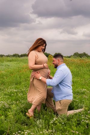 A man kneels in a flower field touching a pregnant woman's belly during an outdoor maternity photoshoot under cloudy skies.