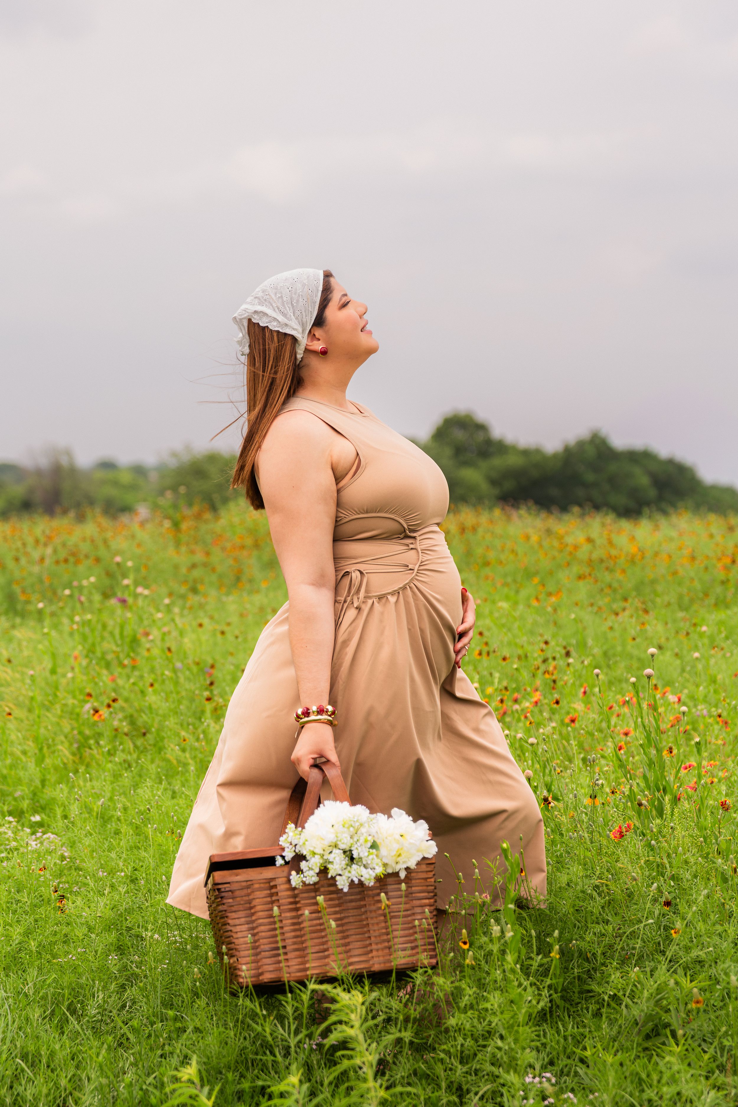 Woman in beige maxi dress holding wicker basket with white flowers standing in colorful wildflower field.