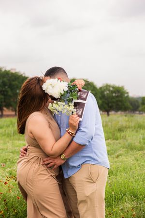 Couple kissing behind white flowers holding ultrasound photo in green field, pregnancy announcement outdoor portrait.