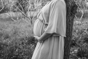 Black and white photo of a pregnant woman in a flowing dress cradling her baby bump outdoors near a tree.