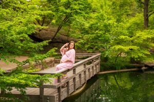 A woman in a pink dress sits on a wooden bridge surrounded by lush green trees reflecting in calm water.