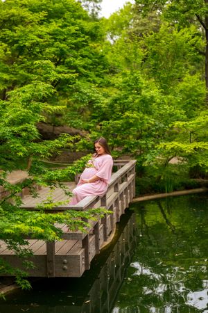 Pregnant woman in pink dress sitting on wooden bridge surrounded by lush green trees reflecting in calm pond water.